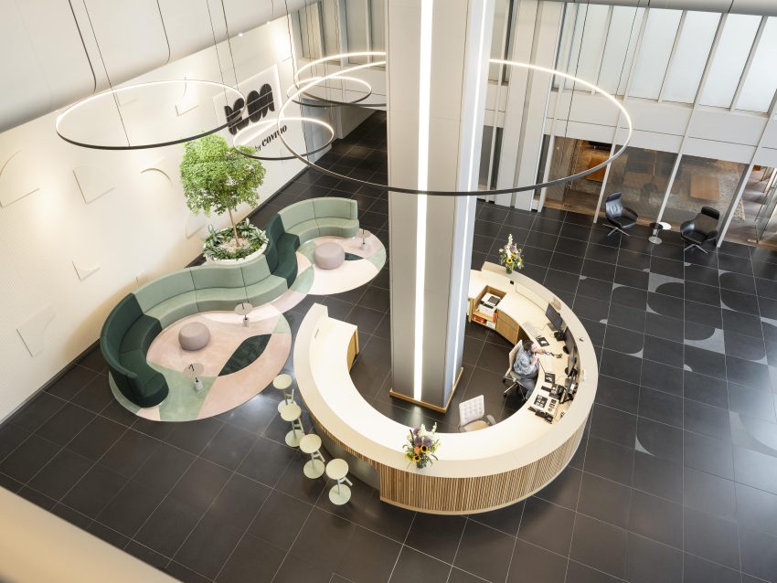 Luftaufnahme eines modernen Empfangsbereichs mit halbkreisförmiger Theke, Sitzbank in Grün- und Rosatönen und großen runden Deckenleuchten