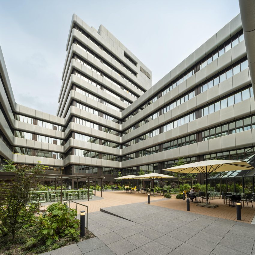 Innenhof mit Sitzbereichen und Sonnenschirmen vor einem mehrstöckigen Bürogebäude mit vielen Fenstern und horizontalen Fassadenelementen