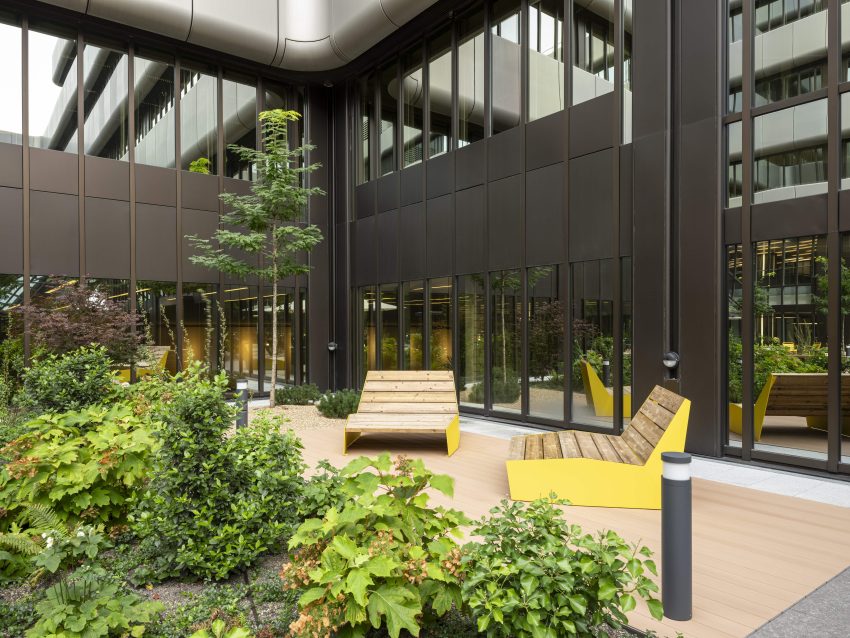 Innenhof mit moderner Fassadenverglasung, Holzliegen mit gelben Metallgestellen und grünen Pflanzen