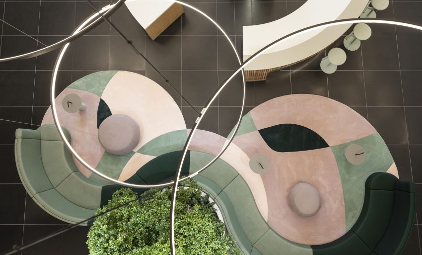 Blick von oben auf geschwungene Sitzmöbel mit pastellfarbenen Teppichen und runden Tischen, darüber geschwungene Leuchten und ein grüner Pflanzenbereich