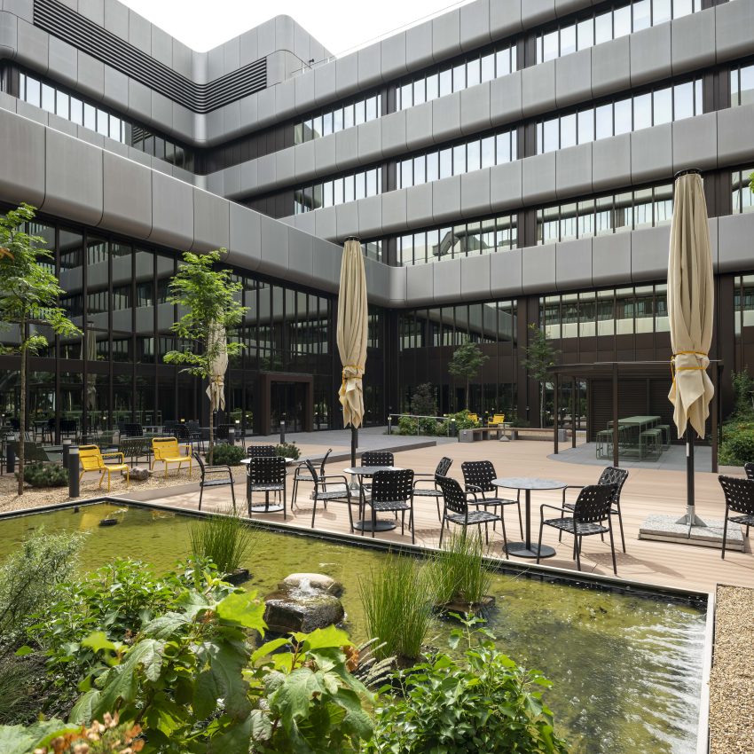 Inner courtyard with pond, seating groups of black chairs and tables, yellow armchairs and closed parasols in front of a modern office building